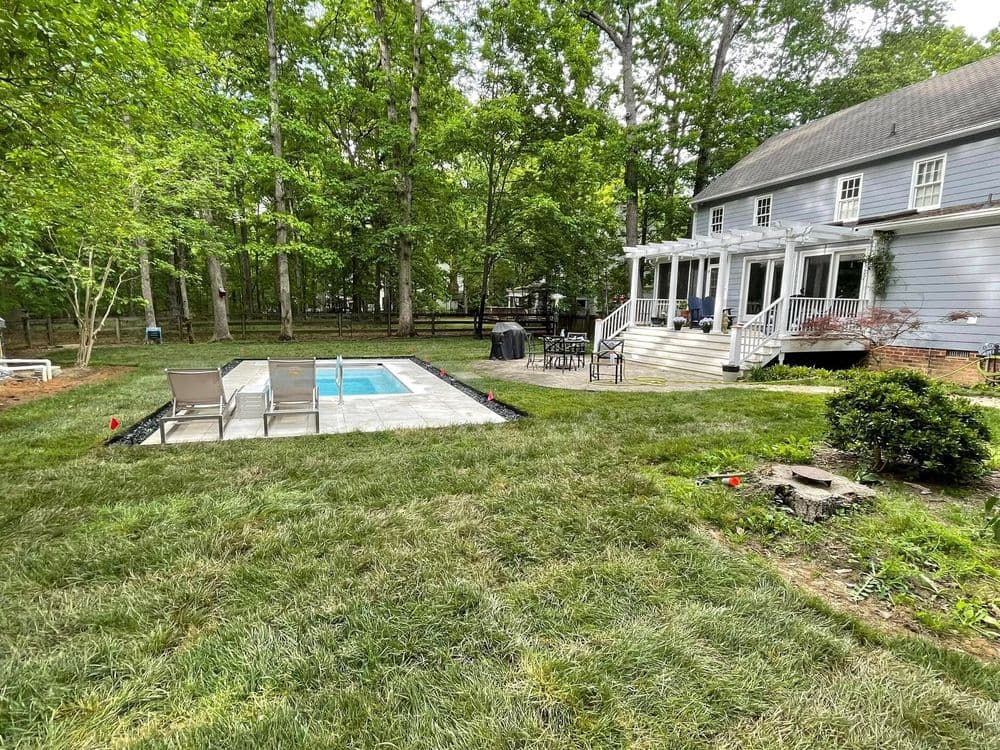 Backyard with a pool, lawn, and a house surrounded by trees and a patio area.