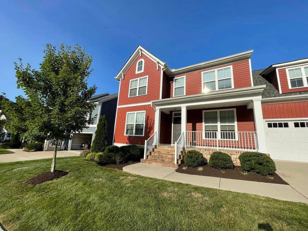 Red two-story house with a porch, neatly landscaped lawn, and blue sky in the background.