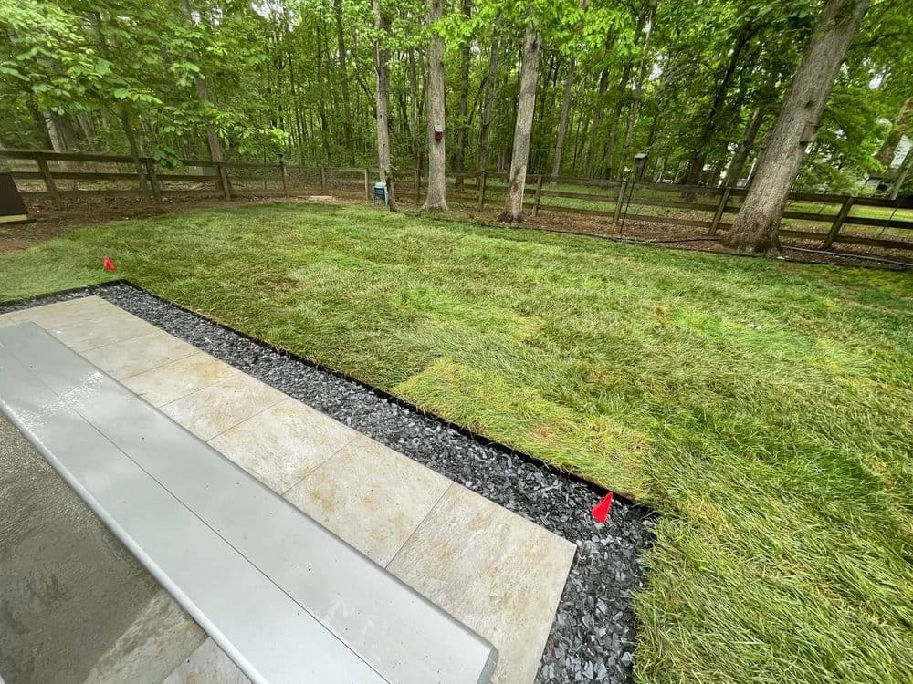 Freshly laid sod in a wooded backyard, bordered by black gravel and seated area.