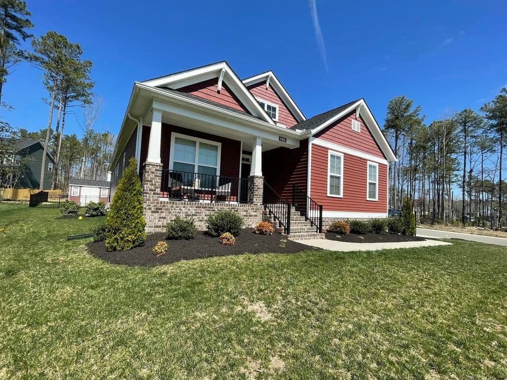 New red and gray house with porch, surrounded by trees and landscaped yard on a sunny day.